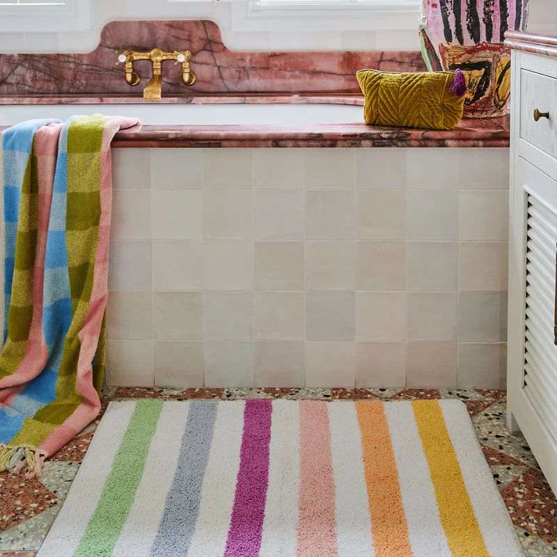 The image shows a colorful bath mat with a striped pattern in various shades. The mat is made from eco-friendly cotton and is part of Kip & Co's Portofino Stripe collection, available at Cove.