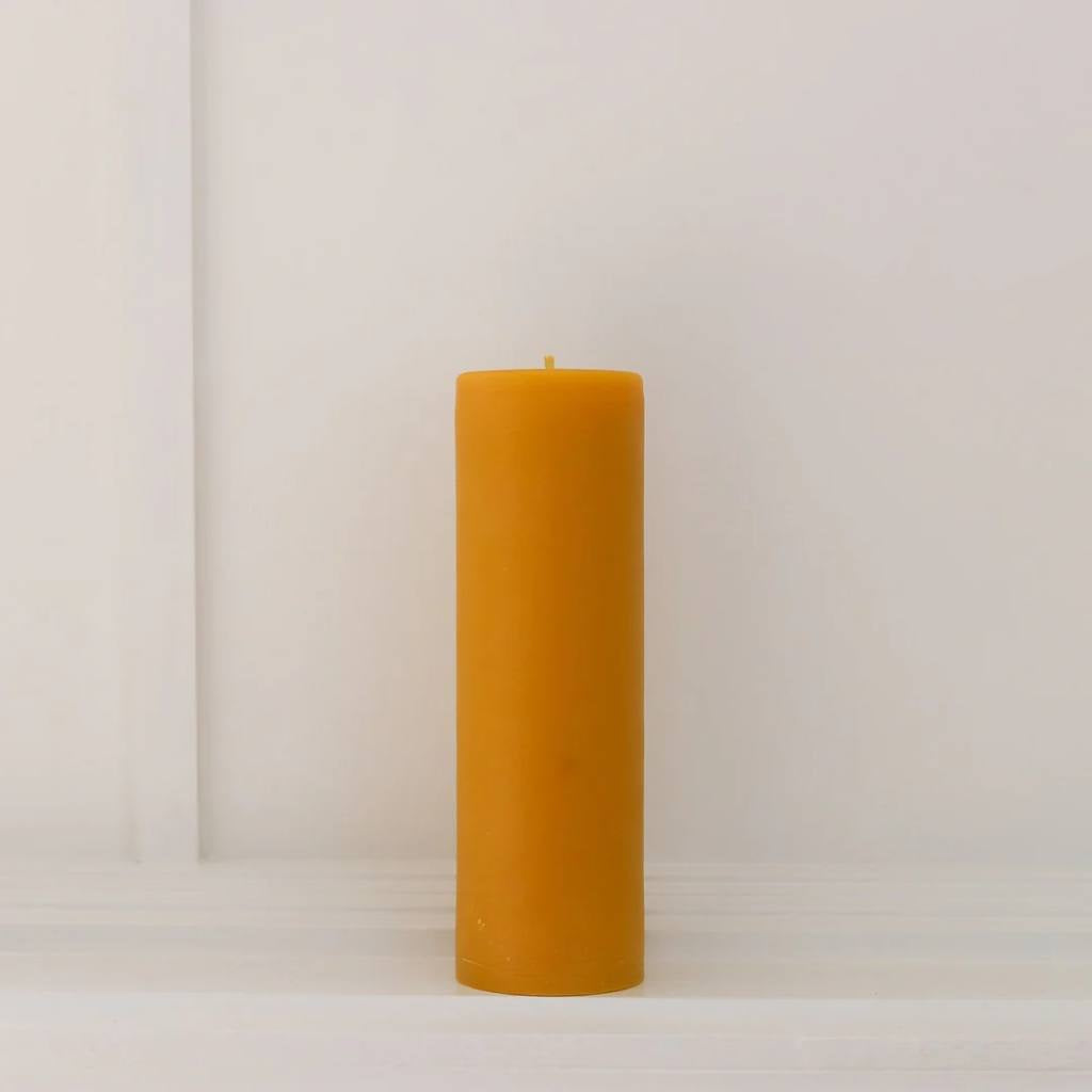 A large, natural-colored pillar candle from The Authentic Bee Co., showcasing its smooth, cylindrical shape and subtle texture. The candle is set against a plain background, highlighting its simplicity and elegance.
