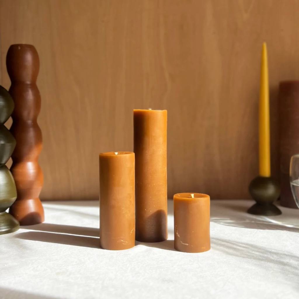 A set of amber-colored pillar candles from The Authentic Honey Co., displayed on a neutral background. The candles vary in height, showcasing their smooth texture and warm, honey-like hue.