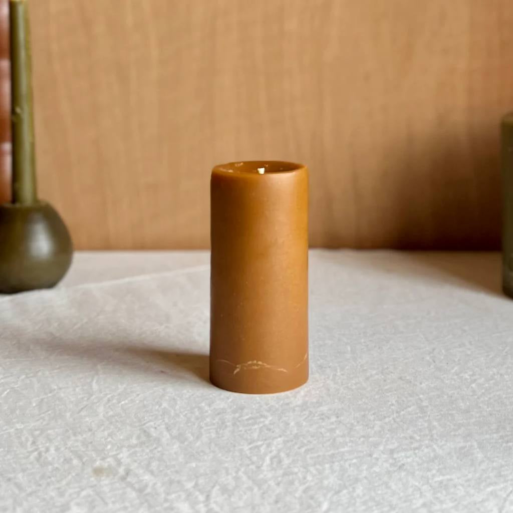 A medium-sized amber pillar candle from The Authentic Honey Co., showcasing a smooth, glossy finish with a warm, honey-like hue. The candle is set against a simple, neutral background, highlighting its rich color and elegant design.