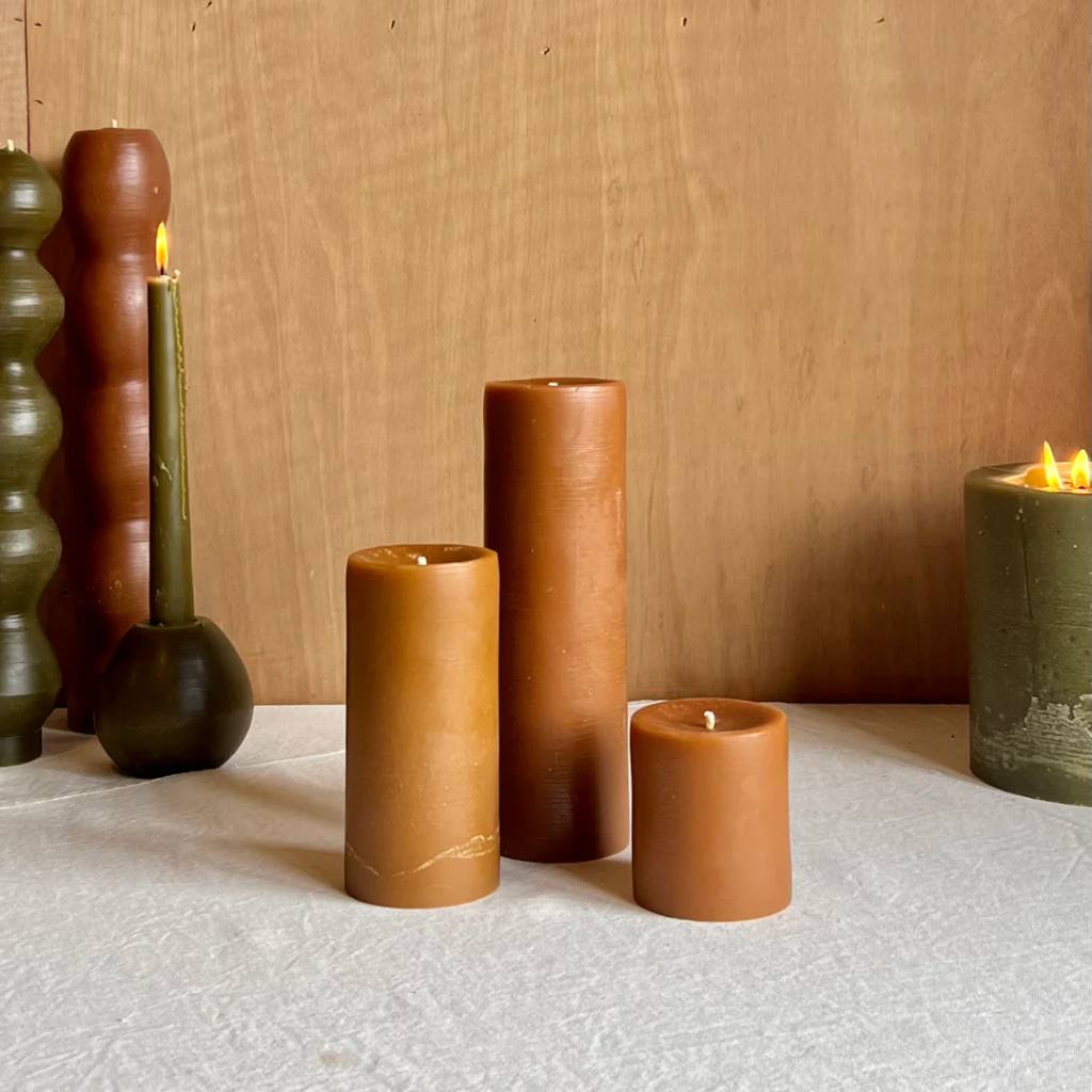 A set of small amber-colored pillar candles from The Authentic Honey Co., displayed on a light wooden surface. The candles have a smooth finish and are arranged in a visually appealing manner, showcasing their warm, honey-like hue.