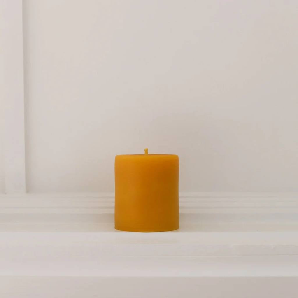 A small, natural beeswax pillar candle from The Authentic Honey Co., showcasing its smooth texture and warm, golden hue. The candle is set against a simple, neutral background, highlighting its handcrafted quality and eco-friendly appeal.