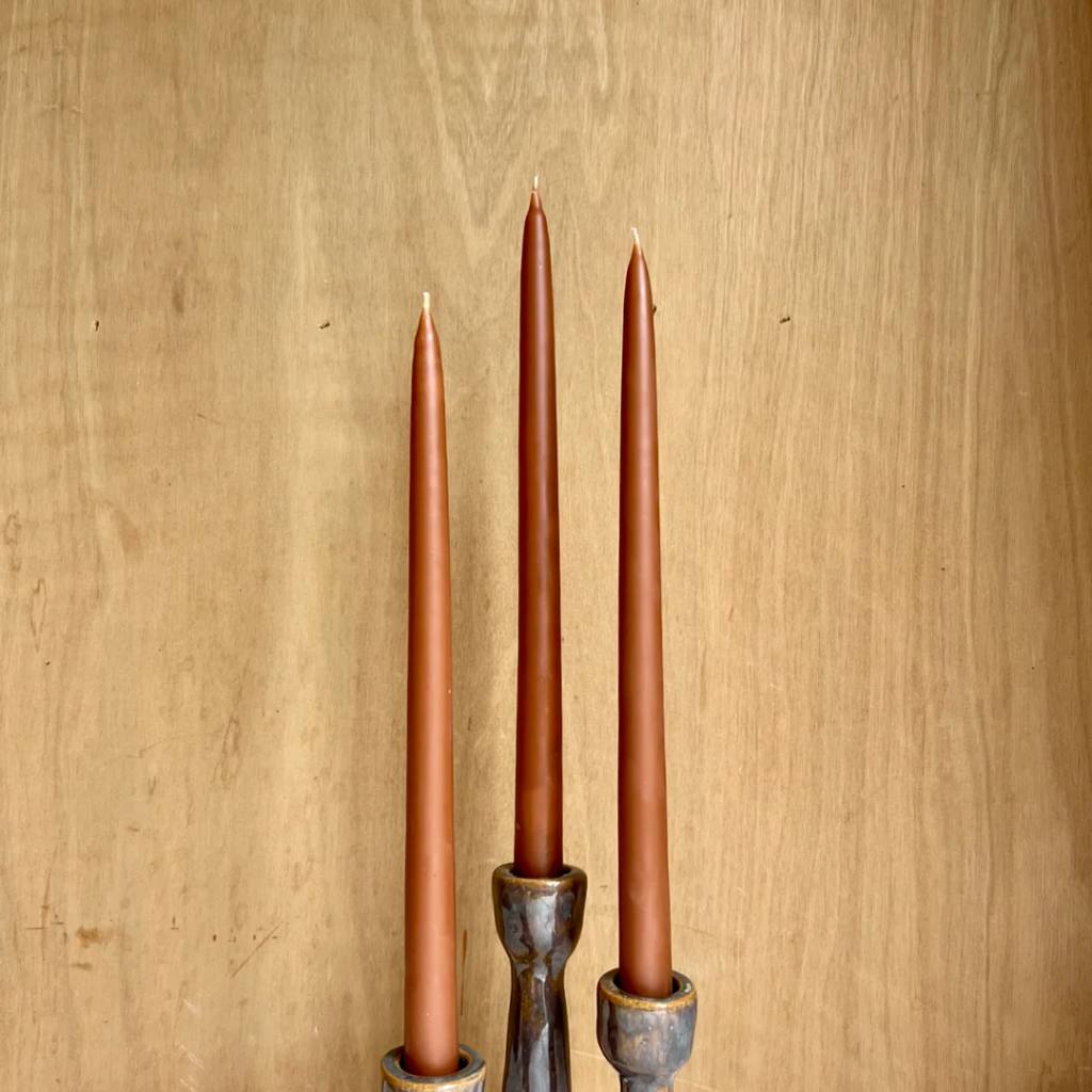 A collection of amber-colored taper candles from The Authentic Honey Co., elegantly displayed on a rustic wooden surface. The candles have a smooth finish and are arranged in a neat row, showcasing their rich, warm hue. The background is softly blurred, highlighting the candles as the focal point of the image.