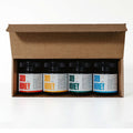 The image shows a Honey Variety Sampler Gift Box containing four different jars of honey. Each jar is neatly arranged in a rectangular box with a clear lid, allowing the golden hues of the honey to be visible. The jars are labeled with different types of honey, showcasing a range of flavors or sources. The packaging is designed to be a gift, with an elegant and appealing presentation.