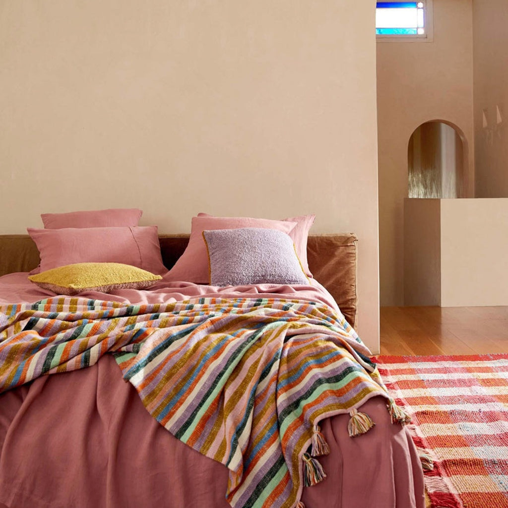 A woven throw blanket with multicolored horizontal stripes draped over a bed, in a room with neutral walls and a small decorative pillow.
