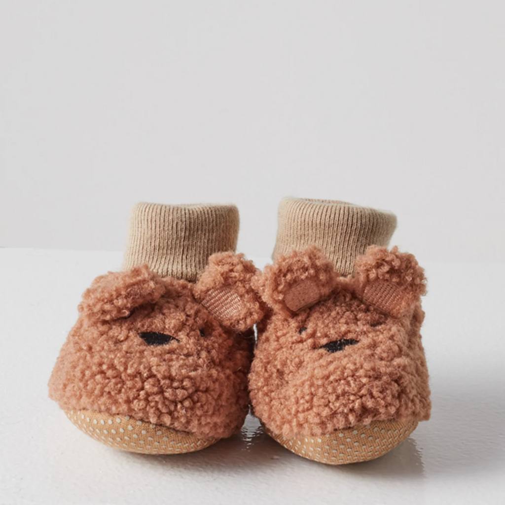 Pair of brown baby booties with pom-pom details on a light gray background