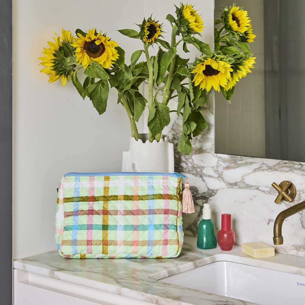 The image is of a quilted cotton toiletry purse from Kip & Co. The purse features a checkered pattern in pastel colors, predominantly mint green and pink, resembling a candy check design. The quilted texture adds a soft, padded appearance to the purse. It has a zipper closure at the top, and the overall look is playful and vibrant, suitable for storing toiletries or small personal items.
