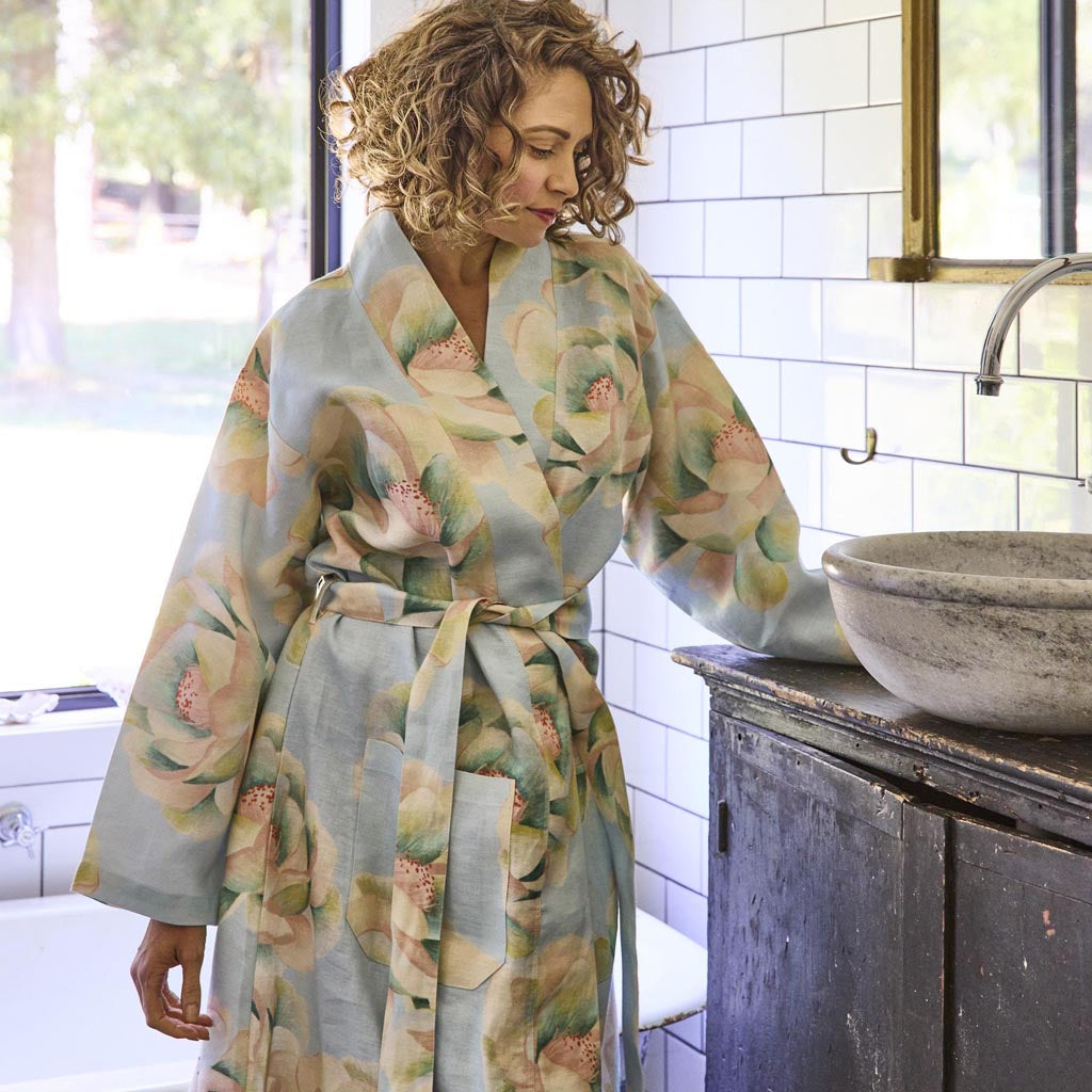 The image shows a blue linen robe with a vibrant floral pattern. The robe features large, colorful flowers in shades of pink, orange, and yellow, with green leaves scattered throughout. It has a relaxed fit with long sleeves and a belt at the waist for tying. The fabric appears lightweight and comfortable, suitable for lounging or casual wear. The robe is displayed against a plain background, highlighting its intricate design and vivid colors.