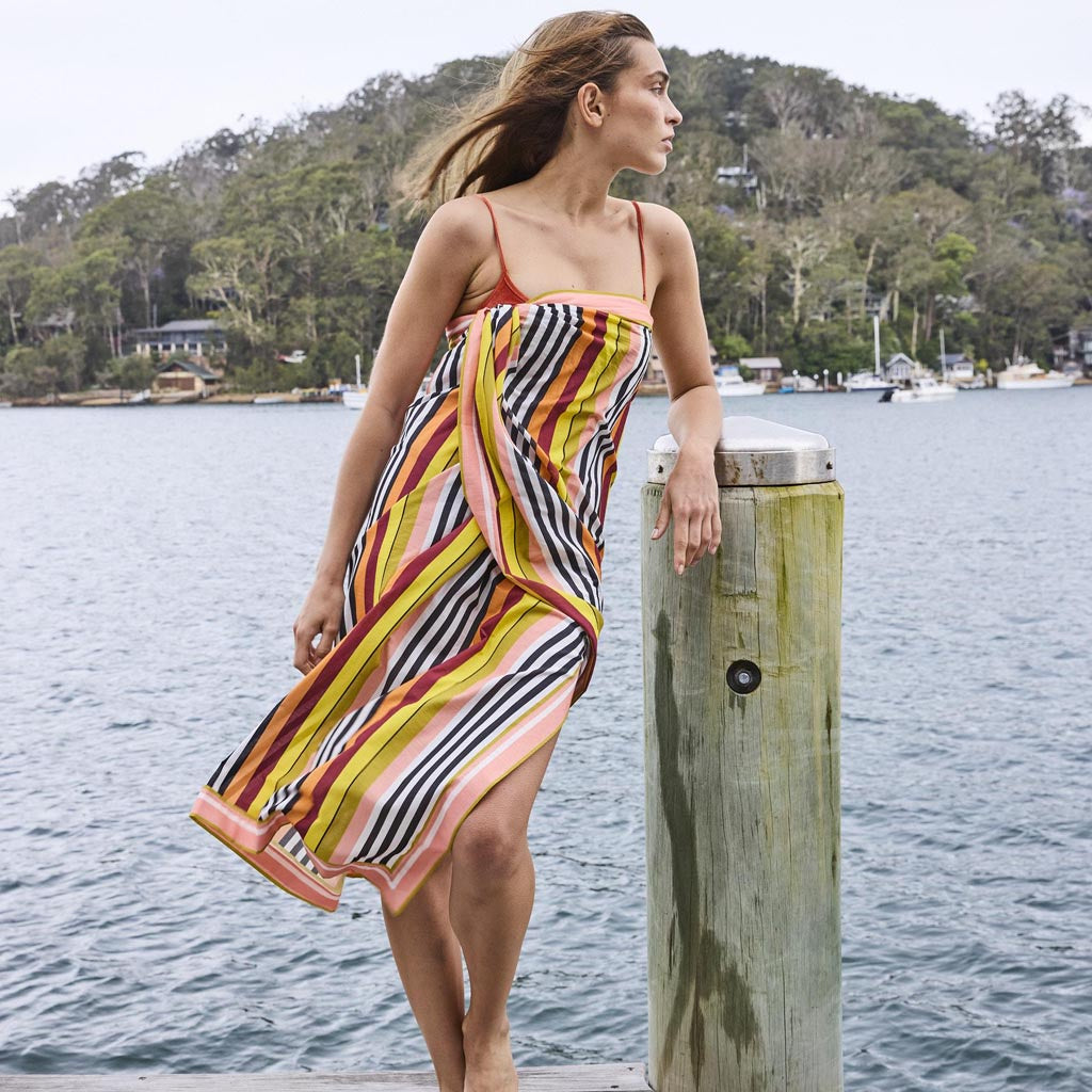 The image is of a colorful striped sarong made from organic cotton. The sarong features a vibrant pattern with horizontal stripes in various colors, including shades of pink, orange, yellow, green, and blue. The fabric appears lightweight and soft, suitable for use as a beach cover-up or a versatile accessory. The sarong is displayed against a plain background, highlighting its bold and lively design.