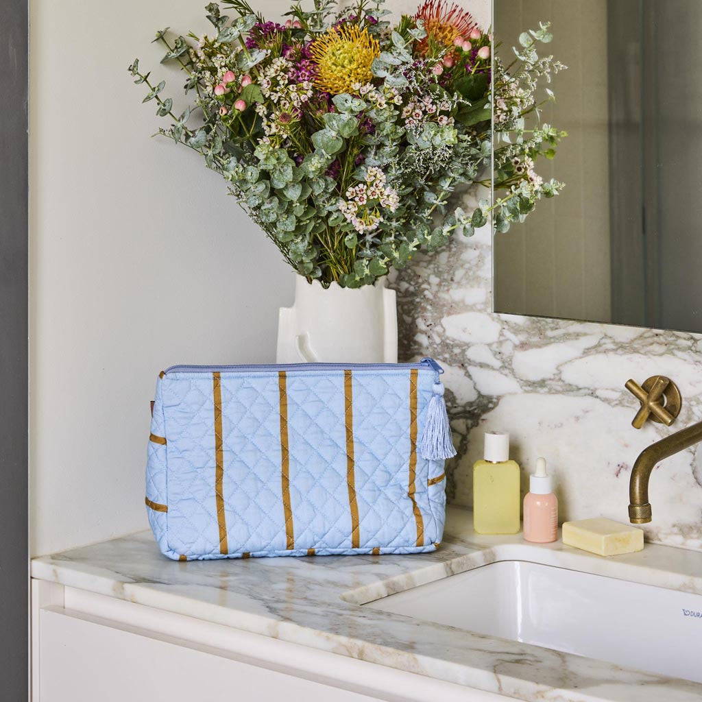 The image shows a quilted cotton toiletry purse with a colorful striped pattern. The stripes are arranged in a variety of colors, including shades of pink, blue, green, and yellow, creating a vibrant and eye-catching design. The purse has a zipper closure at the top, and the quilting gives it a textured appearance. The overall shape is rectangular, and it appears to be a medium-sized accessory suitable for storing toiletries or small personal items.