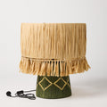 The image shows a stylish table lamp with a unique design. The lamp features a base made of natural raffia material, giving it a textured and organic appearance. The raffia is woven in a way that creates a layered, almost basket-like effect. The lampshade is simple and complements the base, likely made of a neutral fabric that allows the focus to remain on the intricate design of the raffia base. The overall look of the lamp is modern yet earthy, making it a versatile piece for various interior styles.