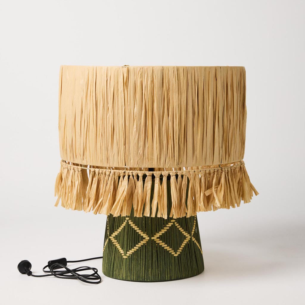 The image shows a stylish table lamp with a unique design. The lamp features a base made of natural raffia material, giving it a textured and organic appearance. The raffia is woven in a way that creates a layered, almost basket-like effect. The lampshade is simple and complements the base, likely made of a neutral fabric that allows the focus to remain on the intricate design of the raffia base. The overall look of the lamp is modern yet earthy, making it a versatile piece for various interior styles.