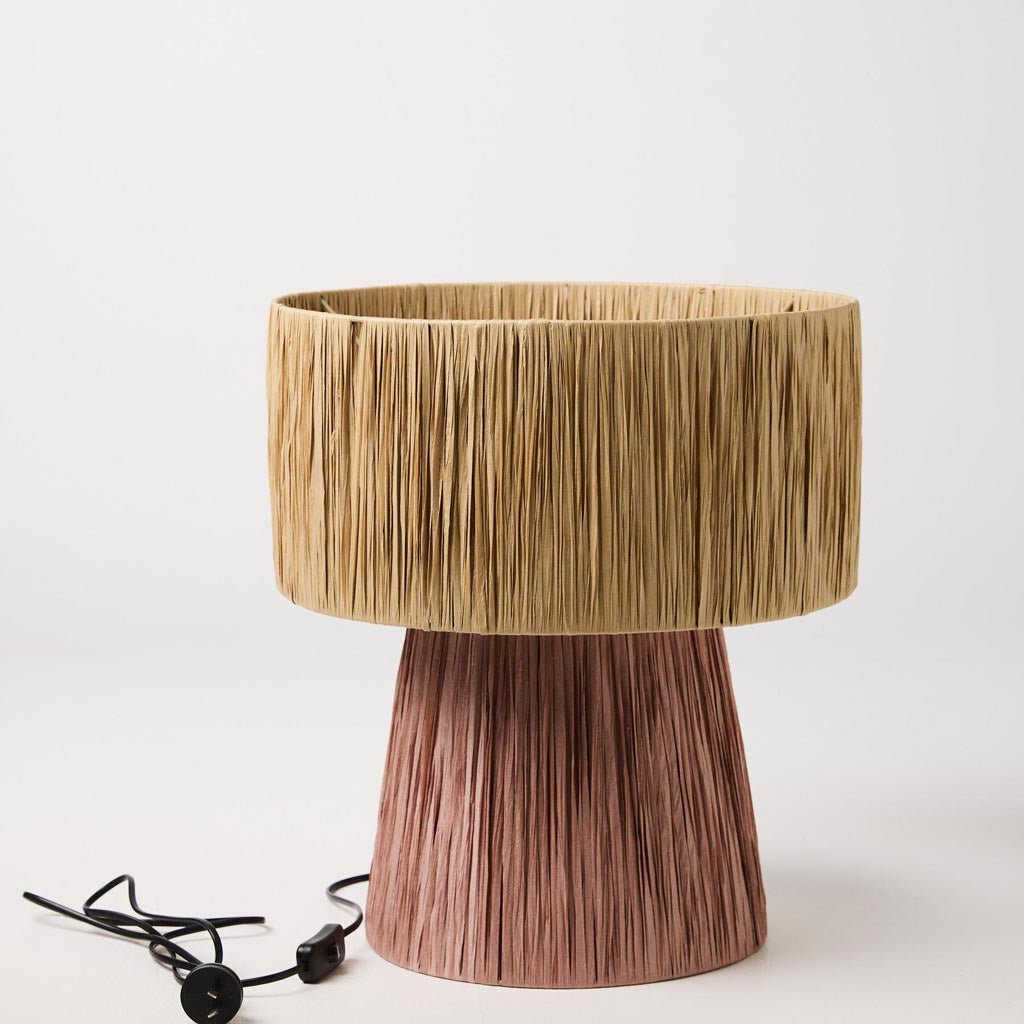 The image is of a decorative lamp made from raffia material. The lamp has a natural, earthy appearance with a woven texture, showcasing the intricate craftsmanship of the raffia fibers. The lampshade is likely in a neutral or light color, complementing the organic feel of the design. The lamp's base may also feature similar natural materials or a simple design to keep the focus on the unique raffia shade. This type of lamp would be suitable for adding a touch of bohemian or rustic charm to a room.