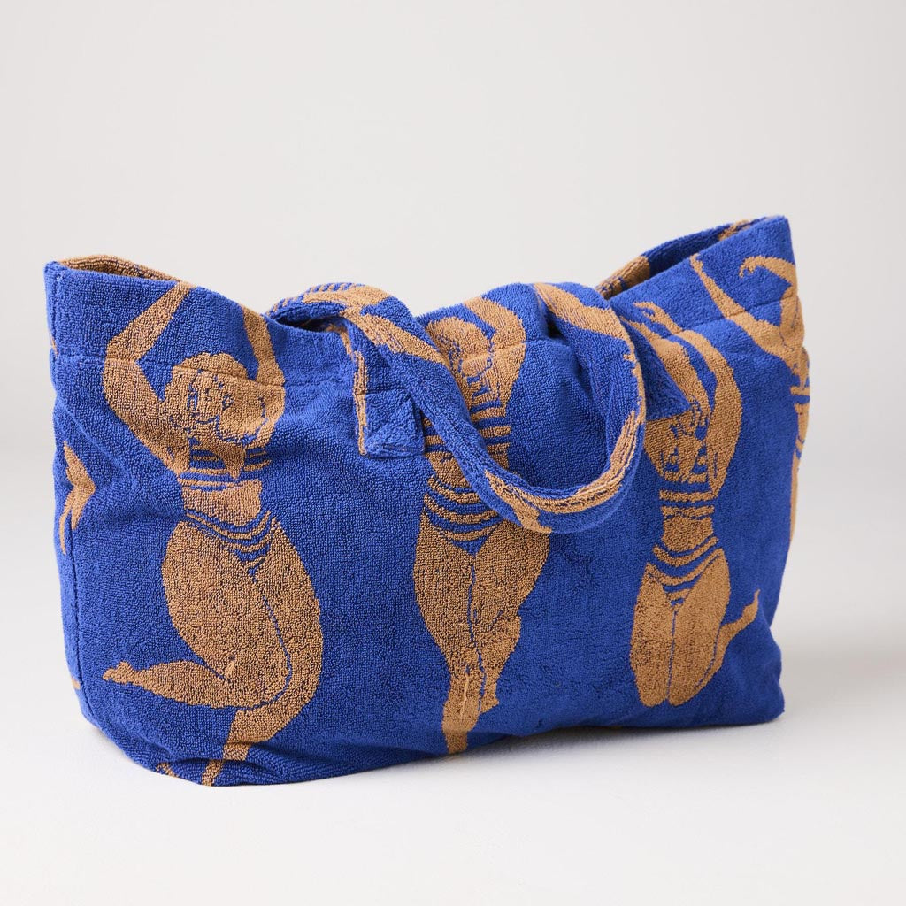 The image shows a large, colorful beach bag made from terry cloth material. The bag features a vibrant, multicolored striped pattern with shades of pink, orange, yellow, and blue, reminiscent of a sunset or a sun-kissed theme. It has sturdy handles for easy carrying and appears spacious, ideal for holding beach essentials. The overall design is cheerful and summery, perfect for a day at the beach or pool.