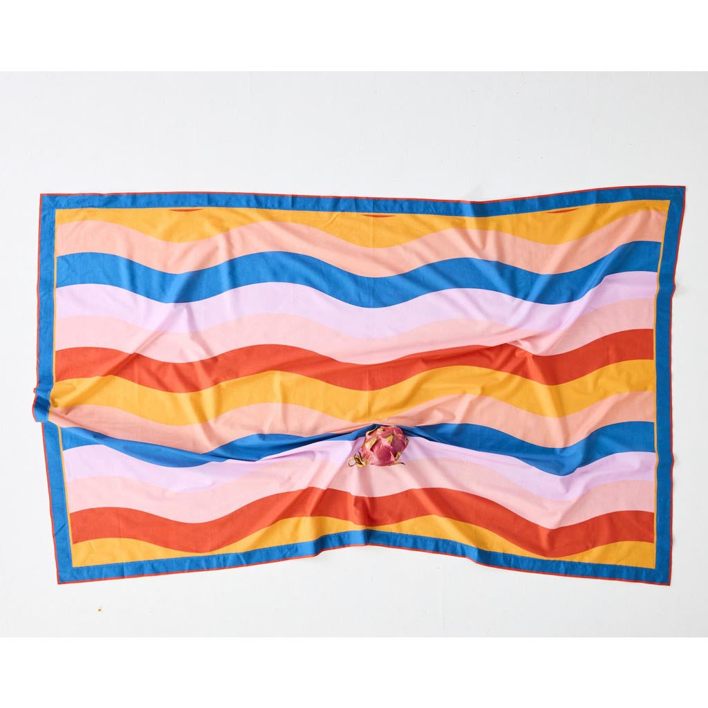 The image is of a colorful sarong made from organic cotton. The design features a vibrant and playful pattern with various shades of blue, green, and yellow, reminiscent of ocean waves or a beach scene. The fabric appears lightweight and soft, suitable for wearing as a cover-up at the beach or pool. The sarong is displayed against a plain background, highlighting its intricate design and vivid colors.