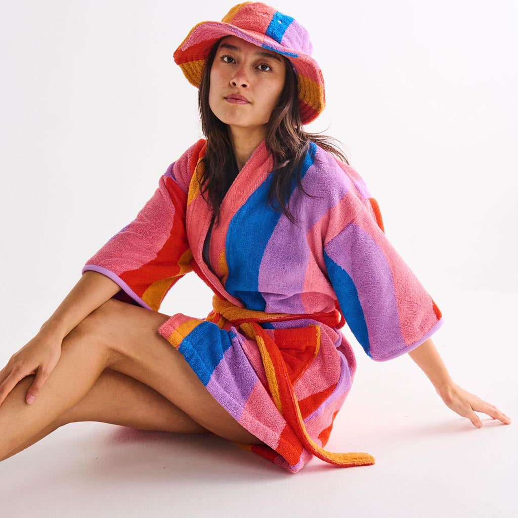 The image is of a children's robe made from terry cloth material. The robe features a colorful and playful design with a surfing theme. It includes various vibrant patterns and illustrations, such as surfboards, waves, and beach-related motifs. The robe has a hood, long sleeves, and a belt for tying around the waist, providing a cozy and comfortable fit for kids. The overall look is fun and suitable for beach or bath time.