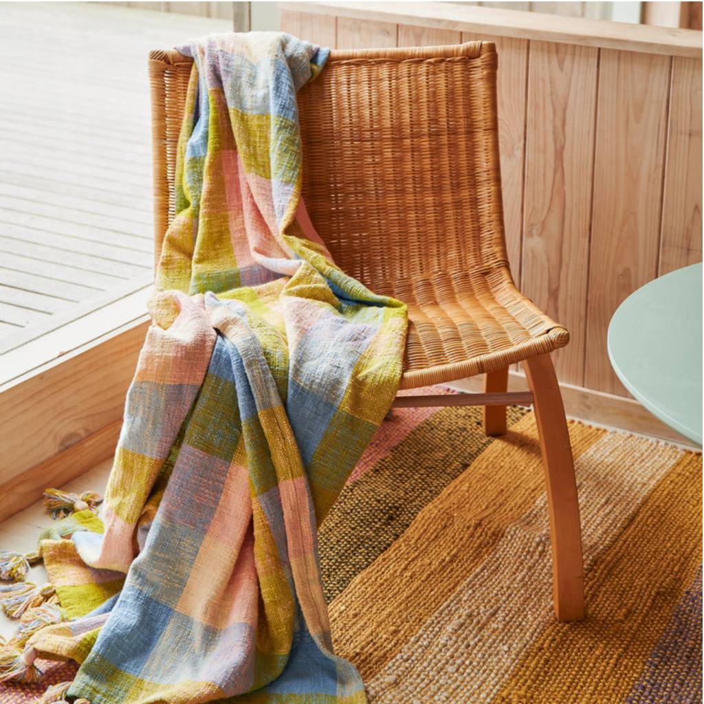 The image likely features Kip & Co's Corfu Tartan Throw, showcasing its vibrant colors and intricate tartan pattern. The throw is made from 100% cotton, adding a cozy and stylish touch to any space.