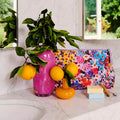The image shows a Kip & Co Forever Floral Lilac Purse, featuring a vibrant floral pattern in shades of lilac and other complementary colors. The purse is designed to be both stylish and eco-friendly, offering ample space for essentials.