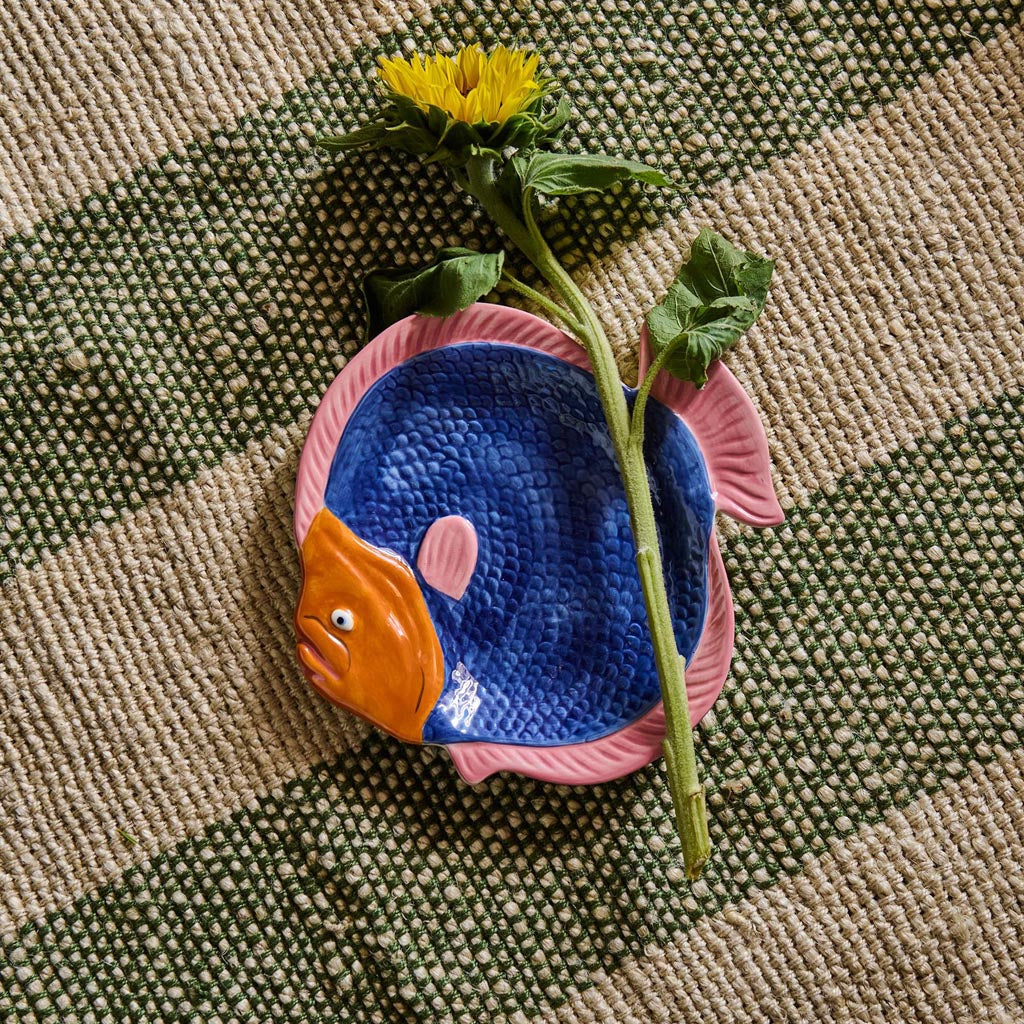 The image shows a colorful, fish-shaped serving dish. The dish is designed to resemble a fish, with intricate details such as scales, fins, and a tail. The body of the fish is painted in vibrant shades of blue, green, and pink, with a pattern that mimics the natural appearance of a fish. The dish is likely made of ceramic or a similar material, giving it a glossy finish. It is placed on a plain background, allowing the vivid colors and unique design to stand out. This serving dish is both decorative and fun