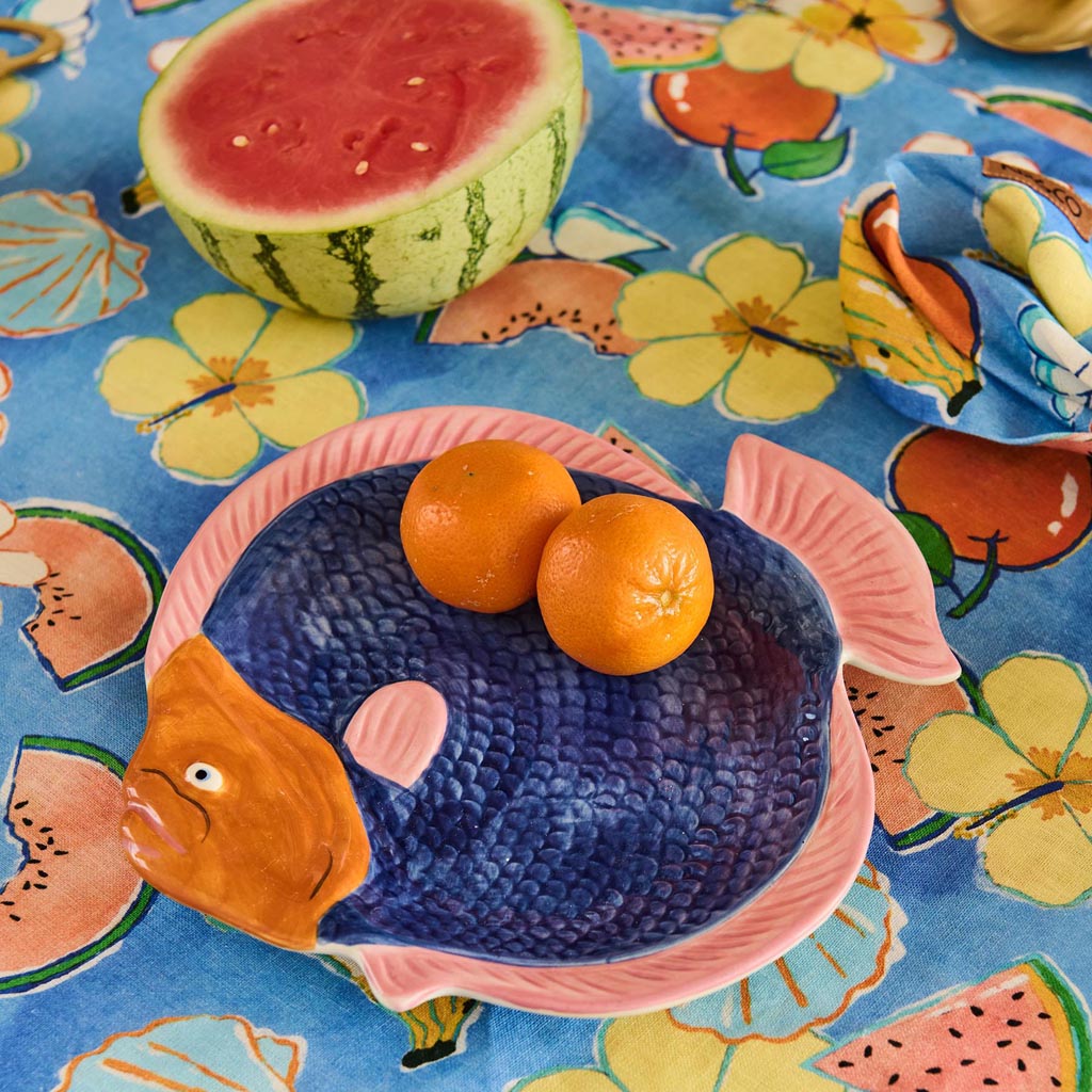 The image is of a colorful serving dish shaped like a fish. The dish features a vibrant design with a mix of bright colors, including shades of blue, pink, yellow, and green, arranged in a pattern that resembles scales. The fish's eye is prominently depicted, adding a playful touch to the overall design. The dish has a glossy finish, enhancing its vivid appearance, and is likely made of ceramic or a similar material. It is set against a plain background, allowing the intricate details and colors of the dish