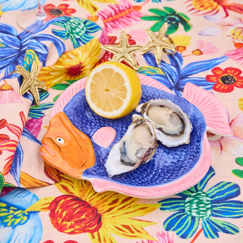 The image shows a colorful, fish-shaped serving dish. The dish is designed to resemble a fish, with intricate details such as scales, fins, and a tail. The colors are vibrant, featuring a mix of blues, greens, and other hues that give it a lively and artistic appearance. The dish is likely made of ceramic or a similar material, and it is placed on a plain background to highlight its design. This serving dish is both functional and decorative, suitable for serving food or as a centerpiece on a dining table.