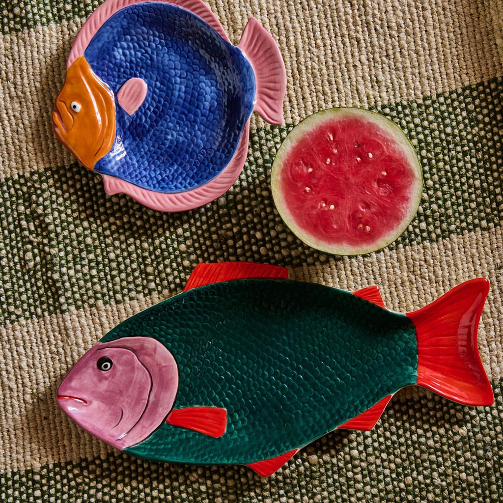 The image shows a colorful, fish-shaped serving dish. The dish is designed to resemble a fish, with intricate details such as scales, fins, and a tail. The colors are vibrant, featuring a mix of blues, greens, and other hues that give it a lively, tropical appearance. The dish is likely made of ceramic or a similar material, and it is placed on a plain background to highlight its design. This serving dish is both decorative and functional, ideal for serving food in a visually appealing way.