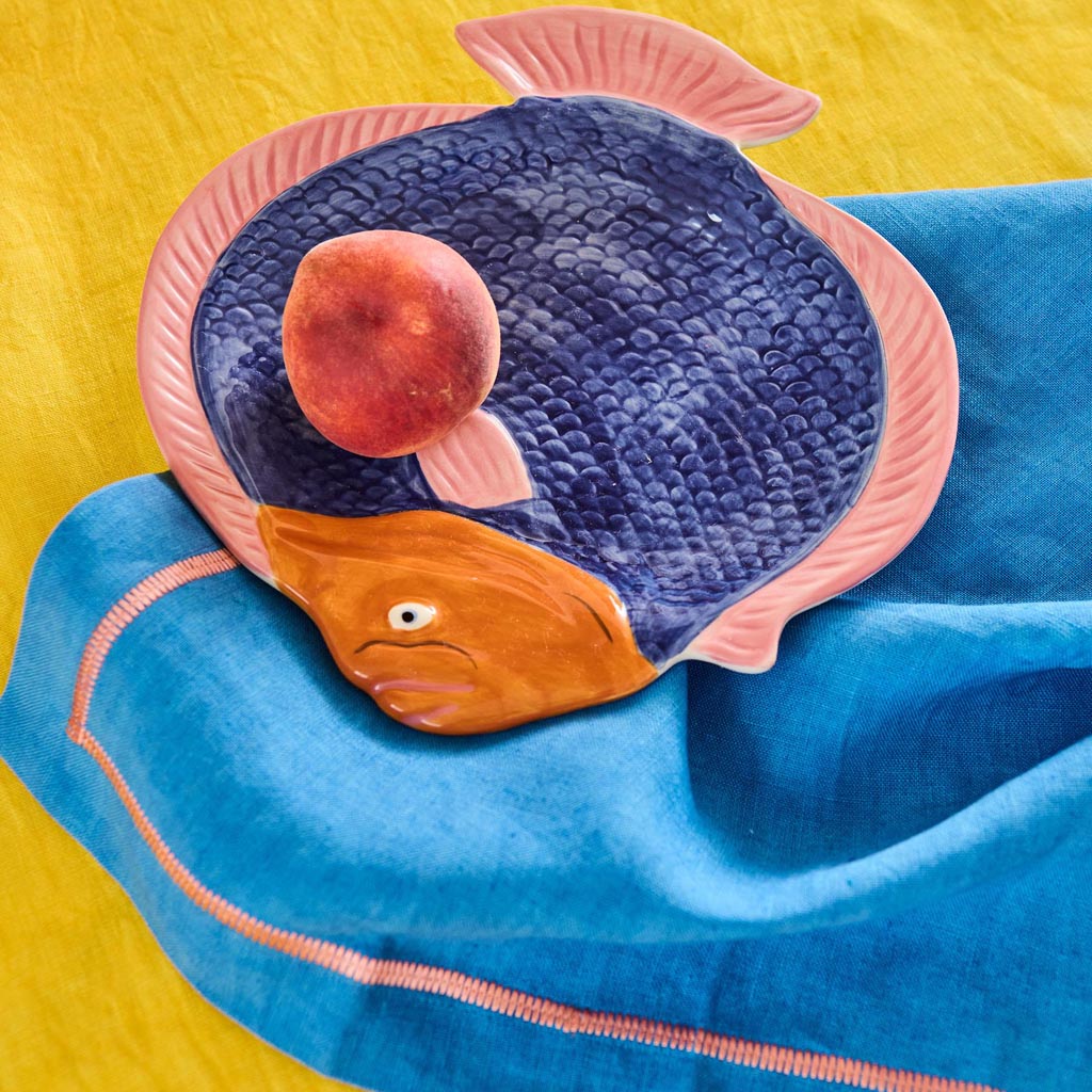 The image is of a colorful serving dish shaped like a fish. The dish features a vibrant design with a mix of bright colors, including shades of blue, pink, yellow, and green, arranged in a pattern that resembles scales. The fish's eye is prominently depicted, adding a playful touch to the overall design. The dish has a glossy finish, enhancing its vivid appearance, and is likely made of ceramic or a similar material. It is set against a plain background, highlighting its intricate and lively design.