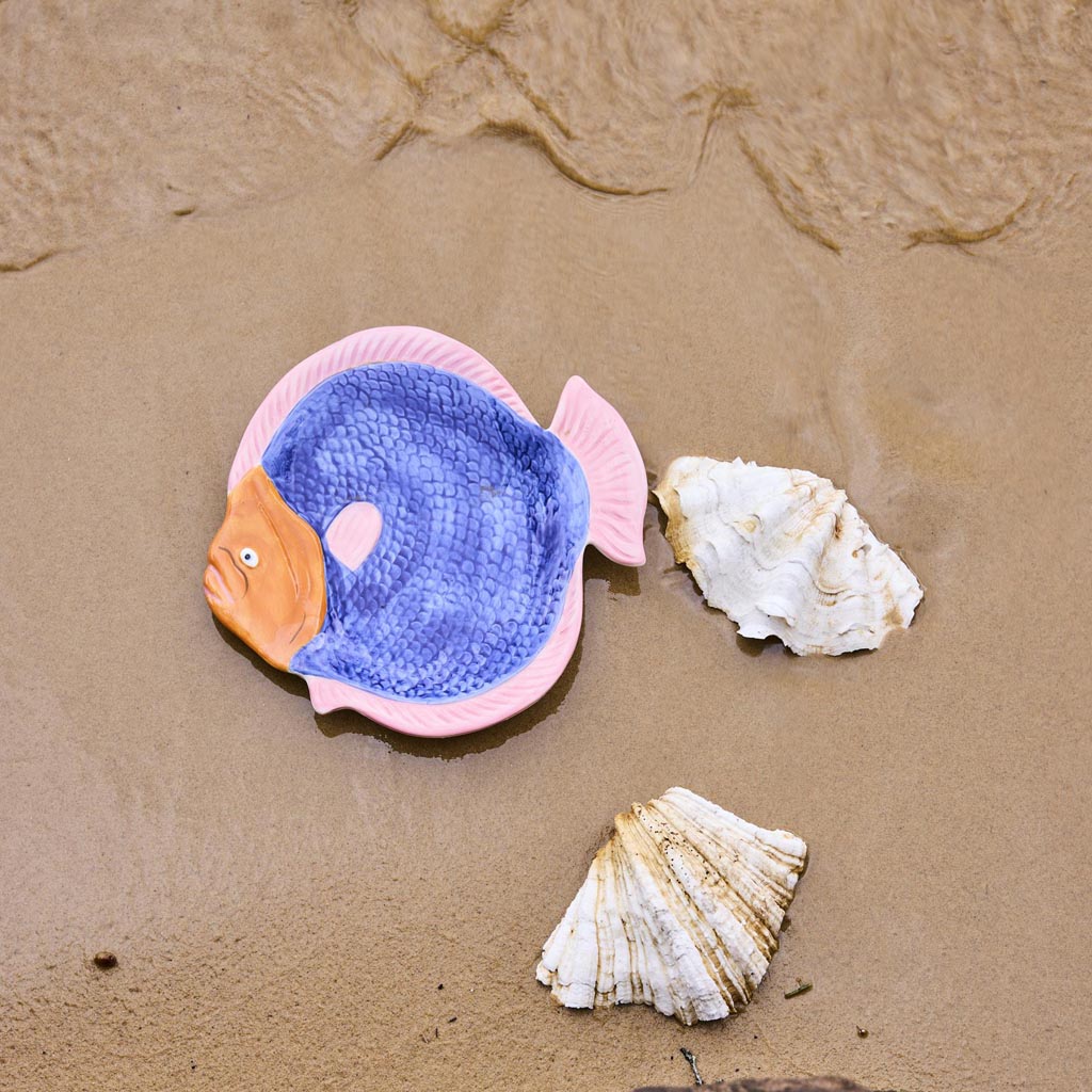 The image is of a colorful, fish-shaped serving dish. The dish is designed to resemble a fish, with a detailed depiction of scales, fins, and a tail. The colors are vibrant, featuring shades of blue, green, and pink, giving it a lively and artistic appearance. The dish is likely made of ceramic or a similar material, and it is intended for serving food, adding a playful and decorative touch to a dining setting.