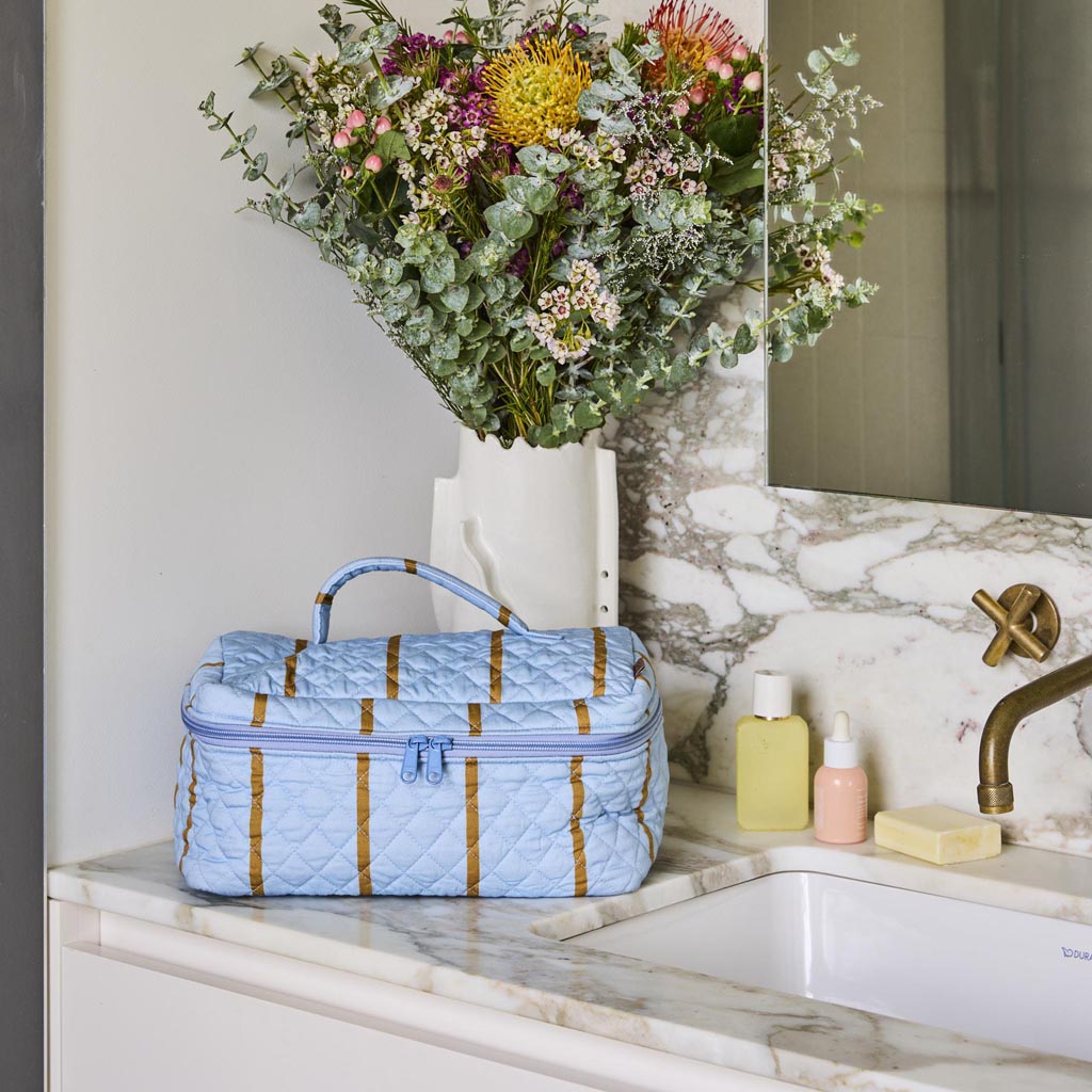 The image shows a quilted cotton toiletry case from Kip & Co. The case features a colorful striped pattern with a variety of vibrant colors, including shades of pink, blue, yellow, and green. The stripes are arranged in a horizontal fashion, giving the case a lively and playful appearance. The quilting adds texture and a sense of depth to the fabric. The case is rectangular in shape and appears to have a zipper closure at the top, making it practical for storing toiletries or other small items. The overall 