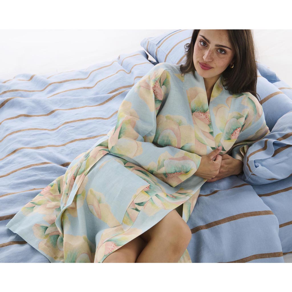 The image is of a blue linen robe with a vibrant floral pattern. The robe features a mix of large, colorful flowers in shades of pink, yellow, and green, creating a bold and eye-catching design. The robe appears to be lightweight and comfortable, with a loose fit and a tie belt at the waist. The background is plain, allowing the intricate details and colors of the robe to stand out prominently.