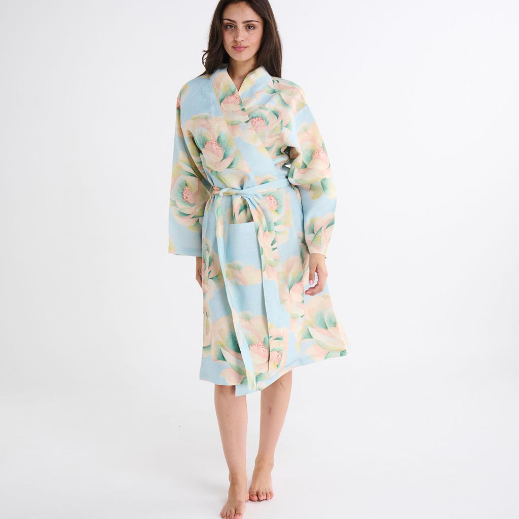 The image shows a blue linen robe with a vibrant floral pattern. The robe features a mix of large, colorful flowers in shades of pink, yellow, and green, creating a bold and eye-catching design. The robe is displayed on a hanger against a plain background, highlighting its flowing fabric and relaxed fit. The sleeves are wide and loose, and the robe includes a matching belt tied around the waist. The overall look is elegant and comfortable, suitable for lounging or as a stylish cover-up.