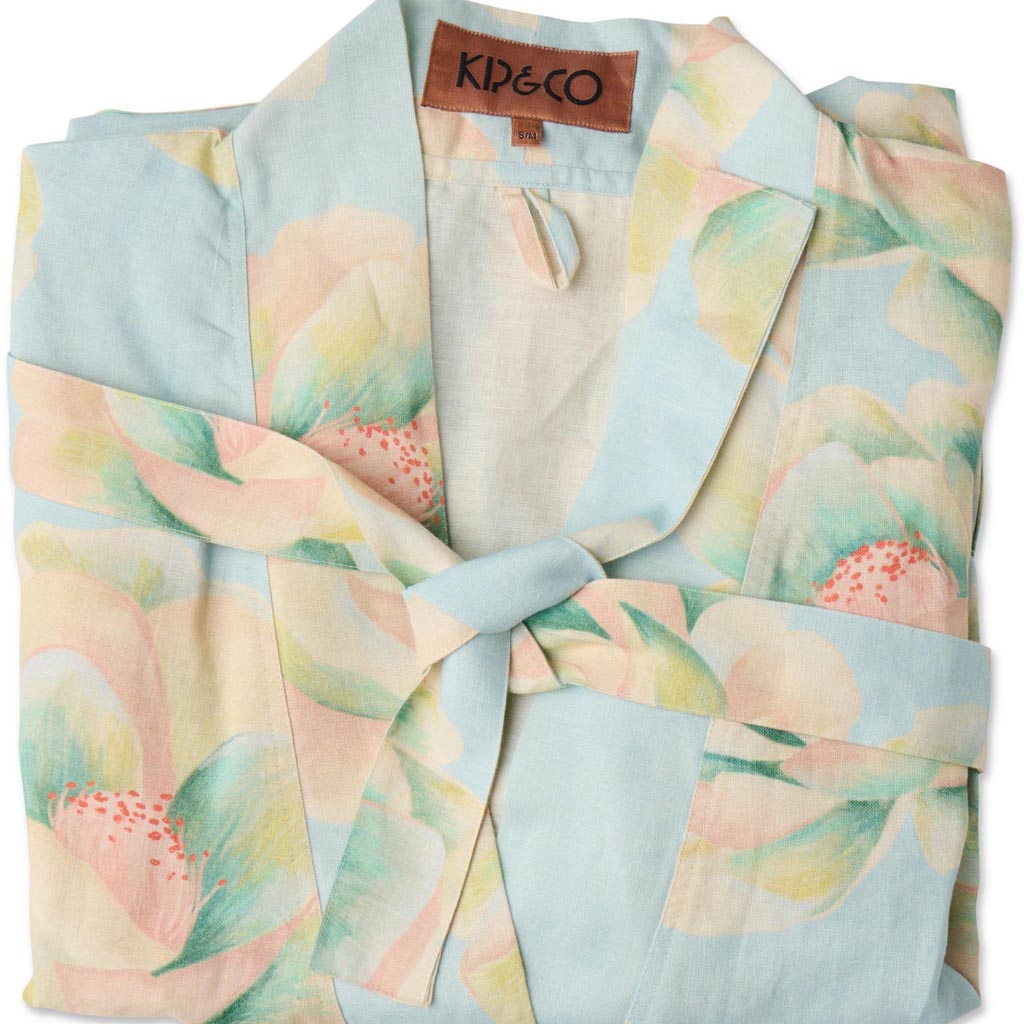 The image shows a blue linen robe with a vibrant floral pattern. The robe features large, colorful flowers in shades of pink, orange, and yellow, with green leaves scattered throughout. It has a relaxed fit with long sleeves and a belt at the waist for tying. The fabric appears lightweight and comfortable, suitable for lounging or casual wear. The robe is displayed against a plain background, highlighting its intricate design and vivid colors.