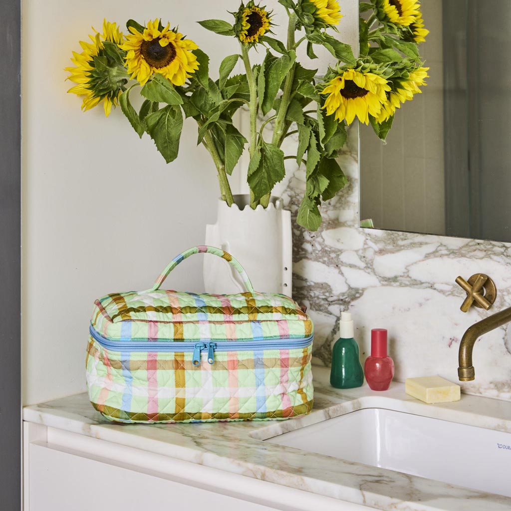 The image is of a quilted cotton toiletry case from Kip & Co. The design features a checkered pattern in pastel colors, primarily mint green and pink, resembling a candy-like appearance. The case has a rectangular shape with a zipper closure on top, and the quilted texture gives it a soft, padded look. The overall aesthetic is playful and vibrant, suitable for storing personal care items.