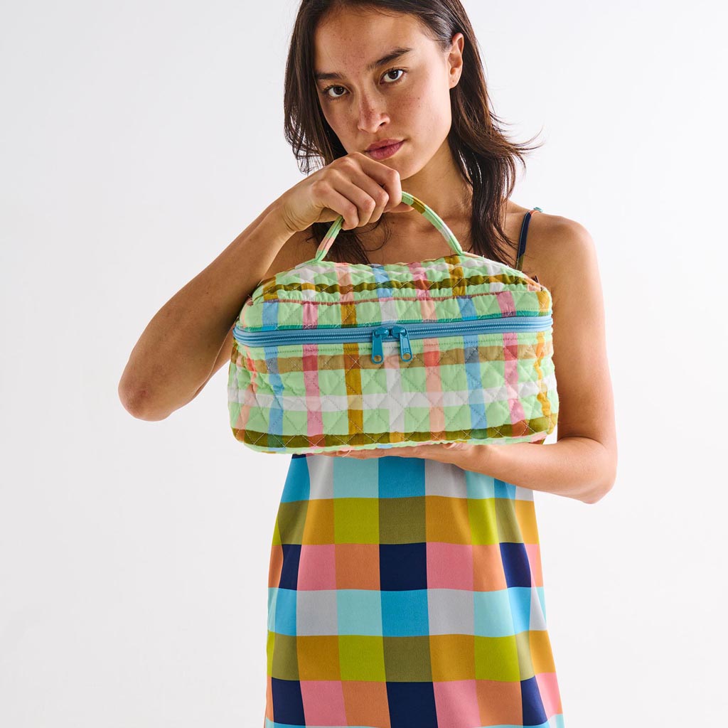 The image is of a quilted cotton toiletry case from Kip & Co. The case features a vibrant and colorful checkered pattern with a mix of pastel colors, predominantly mint green and candy pink. The quilted texture adds a plush, padded appearance to the case. It has a zipper closure at the top, and the overall design is both playful and stylish, suitable for organizing toiletries or small personal items.