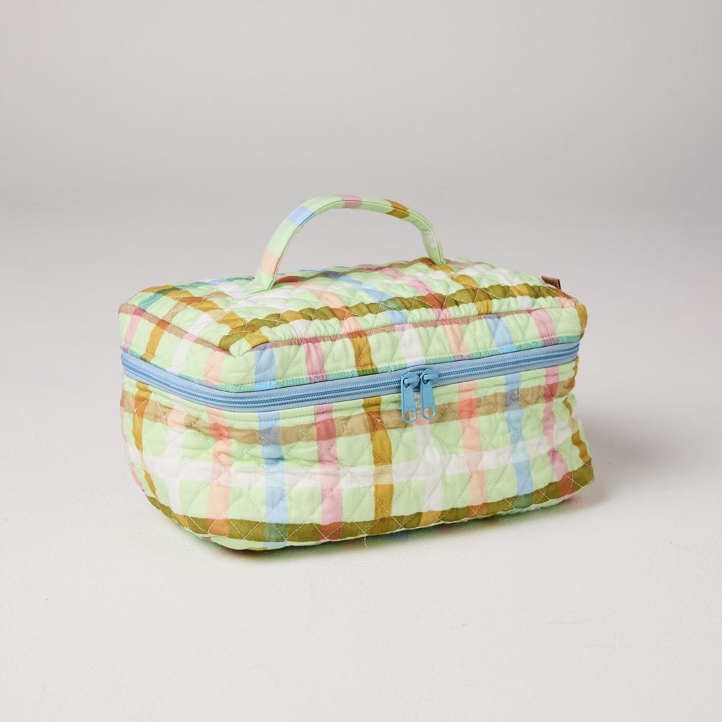 The image shows a quilted cotton toiletry case with a colorful checkered pattern. The pattern consists of pastel colors, including shades of pink, mint green, and yellow, arranged in a grid-like design. The case has a rectangular shape with a zipper closure on the top. The quilting gives it a textured appearance, adding a touch of elegance and style. The overall look is vibrant and playful, making it a fashionable accessory for storing toiletries.