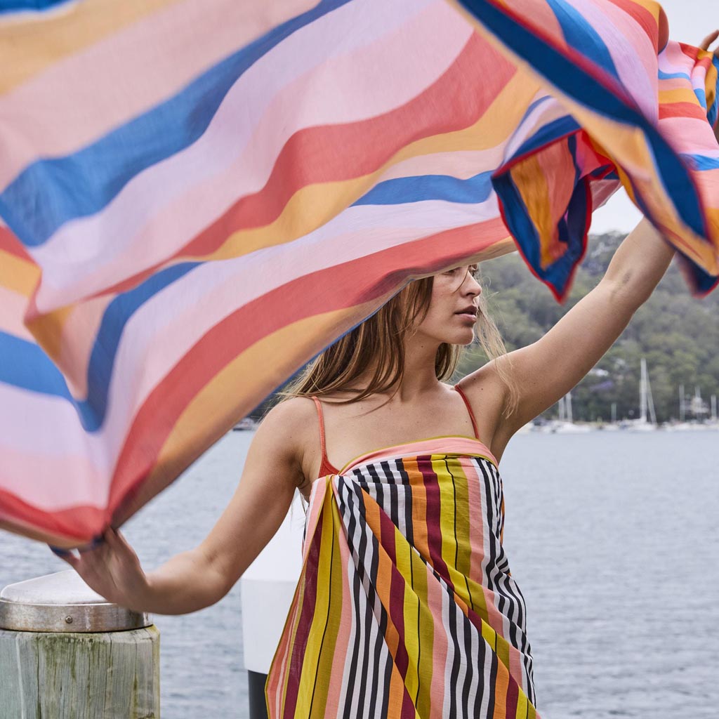 The image is of a colorful striped sarong made from organic cotton. The sarong features a pattern of horizontal stripes in various vibrant colors, including shades of pink, orange, yellow, green, and blue. The fabric appears lightweight and soft, suitable for use as a beach cover-up or a versatile accessory. The sarong is displayed against a plain background, highlighting its bold and lively design.