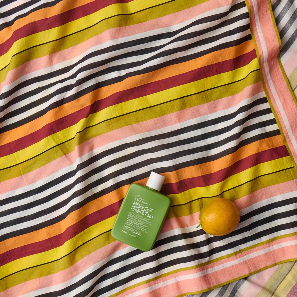 The image shows a colorful striped sarong made from organic cotton. The sarong features a pattern of horizontal stripes in various vibrant colors, including shades of pink, orange, yellow, green, and blue. The fabric appears lightweight and soft, suitable for use as a beach cover-up or a versatile accessory. The sarong is displayed against a plain background, highlighting its bold and lively design.