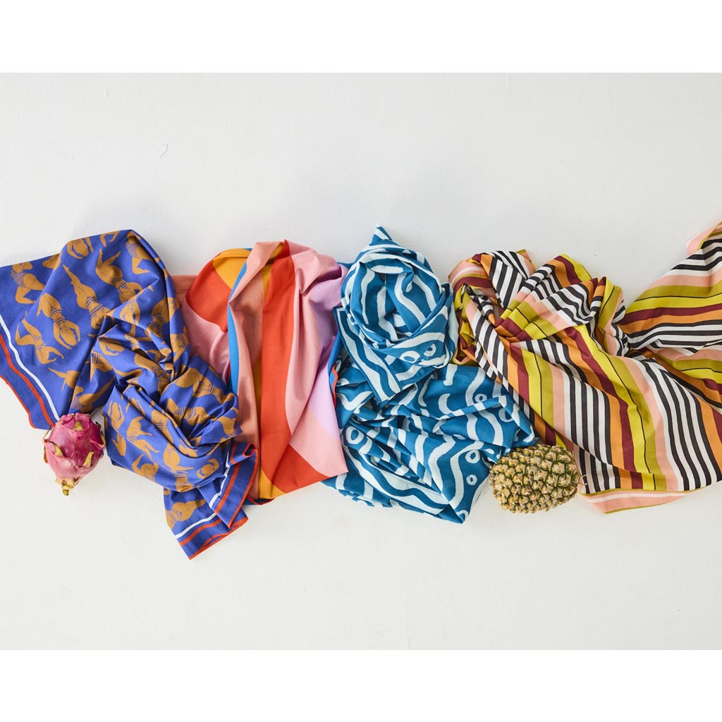 The image shows a colorful striped sarong made from organic cotton. The sarong features a pattern of horizontal stripes in various vibrant colors, including shades of pink, orange, yellow, green, and blue. The fabric appears lightweight and soft, suitable for use as a beach cover-up or a versatile accessory. The background is plain, allowing the sarong's vivid colors to stand out prominently.