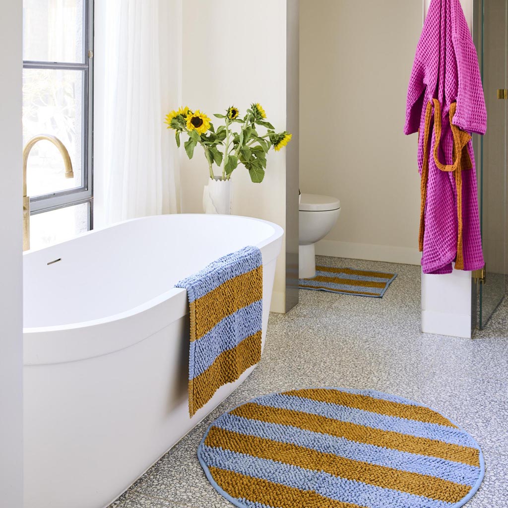 The image is of a colorful bath mat with a striped pattern. The stripes are arranged in a horizontal fashion and feature a variety of vibrant colors, including shades of pink, orange, yellow, green, blue, and purple. The texture of the mat appears soft and plush, suggesting it is made from a comfortable, absorbent material suitable for use in a bathroom setting. The overall design is lively and eye-catching, adding a pop of color to any bathroom decor.