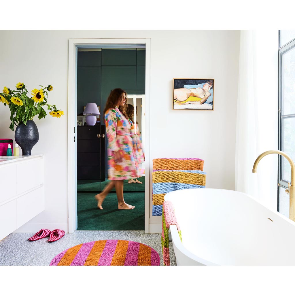 The image is of a colorful bath mat with a striped pattern. The stripes are arranged in a horizontal fashion and feature a variety of vibrant colors, including shades of pink, orange, yellow, green, blue, and purple. The texture of the mat appears soft and plush, suggesting it is made from a comfortable and absorbent material. The overall design is lively and eye-catching, likely intended to add a pop of color to a bathroom setting.