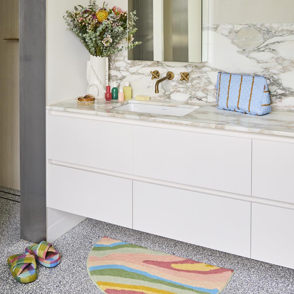 The image shows a quilted cotton toiletry purse with a colorful striped pattern. The stripes are arranged in a variety of colors, including shades of pink, orange, yellow, green, and blue, creating a vibrant and eye-catching design. The purse has a rectangular shape with a zipper closure at the top, and the quilted texture adds a soft, padded appearance. The overall look is playful and stylish, suitable for organizing toiletries or small personal items.