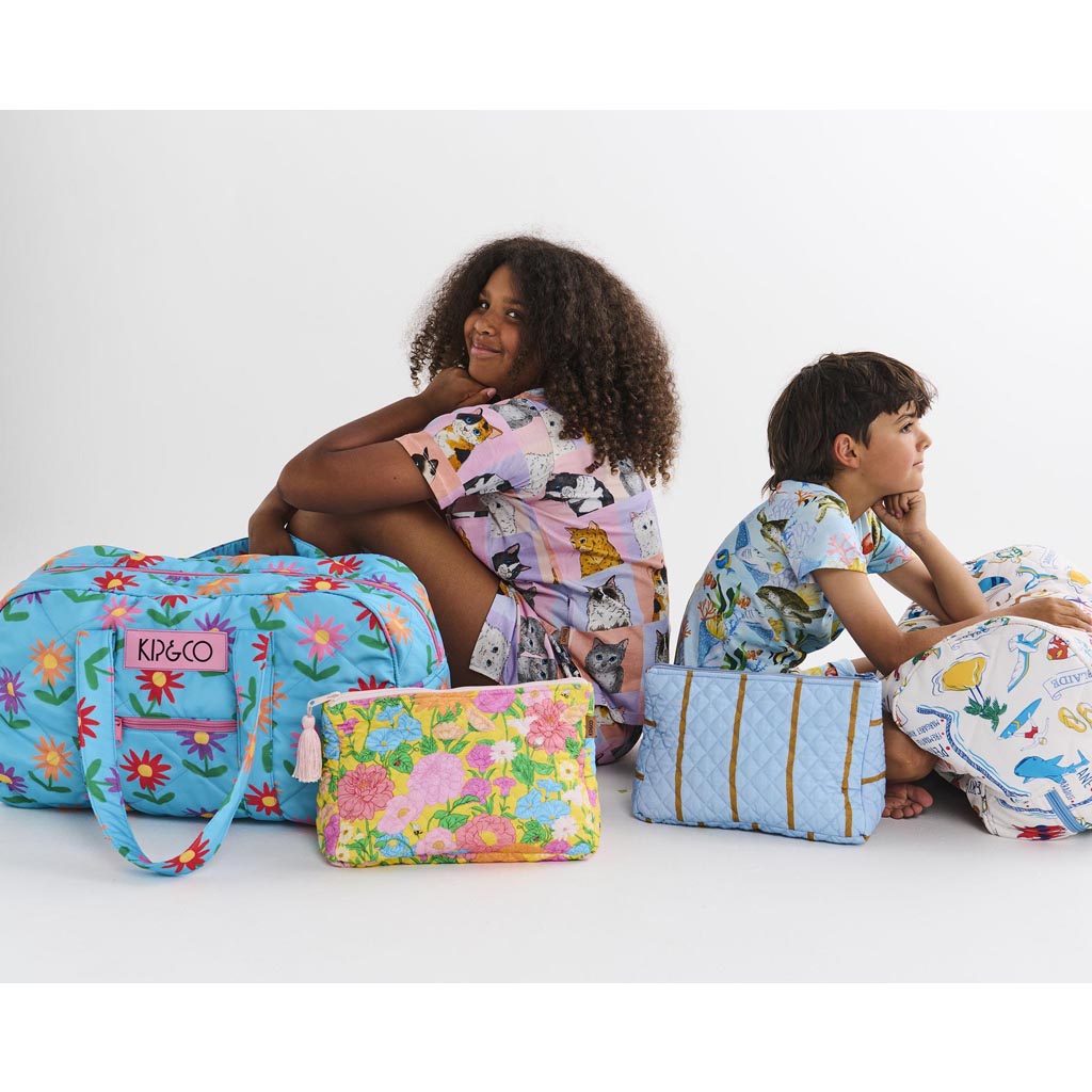 The image is of a quilted cotton toiletry purse from Kip & Co, featuring a colorful striped pattern called "Como Stripe." The purse has a rectangular shape with a zipper closure at the top. The stripes are in various vibrant colors, creating a lively and eye-catching design. The quilted texture adds a layer of dimension and softness to the purse. The background is plain, allowing the purse to be the focal point of the image.