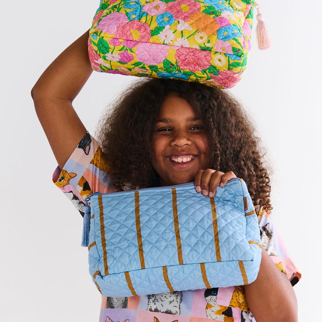 The image is of a quilted cotton toiletry purse from Kip & Co, featuring a colorful striped pattern called "Como Stripe." The purse has a rectangular shape with a zipper closure at the top. The stripes are in various vibrant colors, creating a lively and eye-catching design. The texture of the quilted fabric is visible, adding a sense of depth and softness to the appearance of the purse.