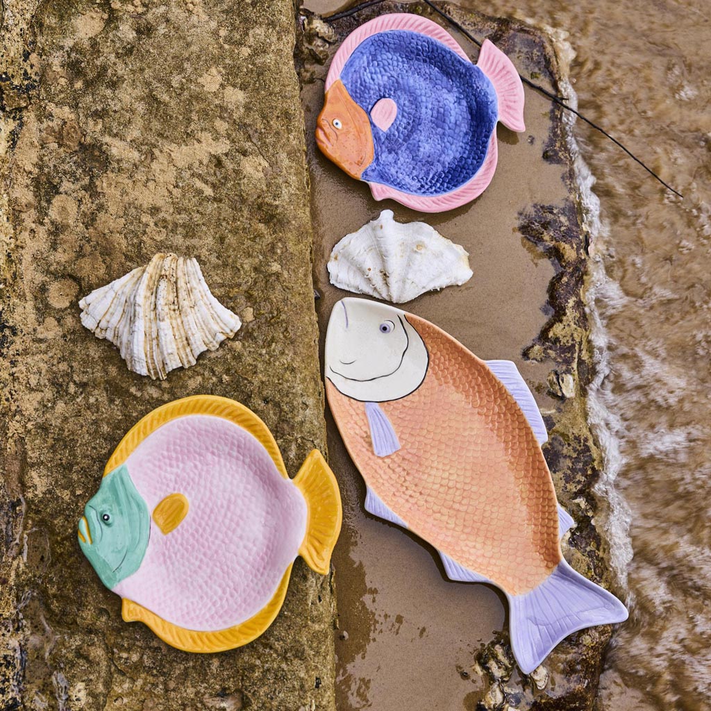 The image shows a decorative serving dish shaped like a fish. The dish is designed with vibrant colors and intricate patterns, resembling a coral reef theme. The body of the fish features a mix of coral pink, blue, and green hues, with detailed scales and fins that add texture and depth. The eye of the fish is prominently designed, giving it a lively appearance. The dish is likely made of ceramic or a similar material, and it is placed on a plain background to highlight its artistic design.