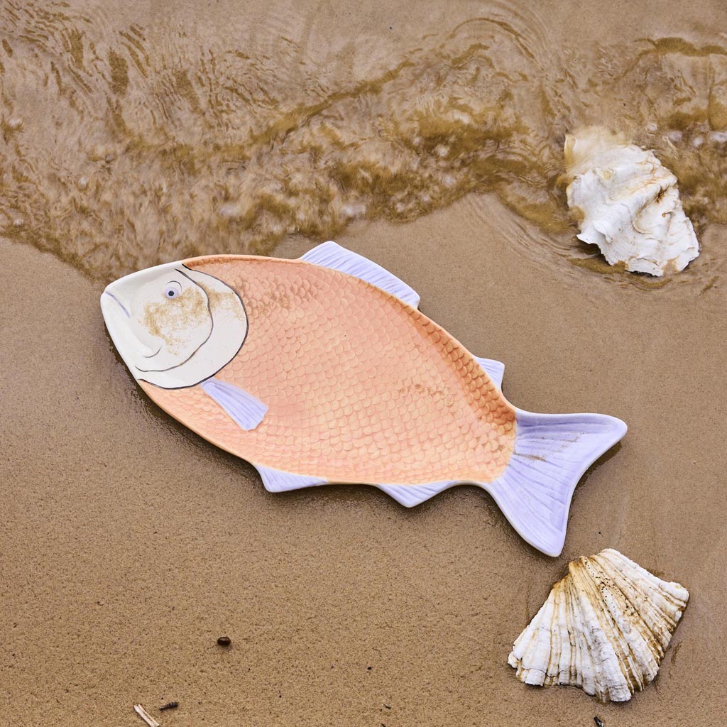 The image shows a decorative serving dish shaped like a fish. The dish is designed with vibrant colors and intricate patterns, reminiscent of tropical or oceanic themes. The body of the fish is adorned with various shades of blue, green, and hints of other colors, creating a lively and eye-catching appearance. The dish is likely made of ceramic or a similar material, and it is intended for serving food, adding a playful and artistic touch to dining settings.