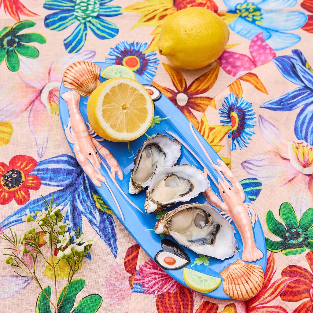 The image is of a large oval platter from Kip & Co, featuring a vibrant and colorful design. The platter is adorned with an artistic depiction of various sea creatures and marine elements, such as fish, shells, and seaweed, all rendered in a playful and whimsical style. The colors are bright and eye-catching, with a mix of blues, greens, pinks, and yellows, creating a lively and cheerful appearance. The platter's design is both functional and decorative, making it suitable for serving food or as a statement