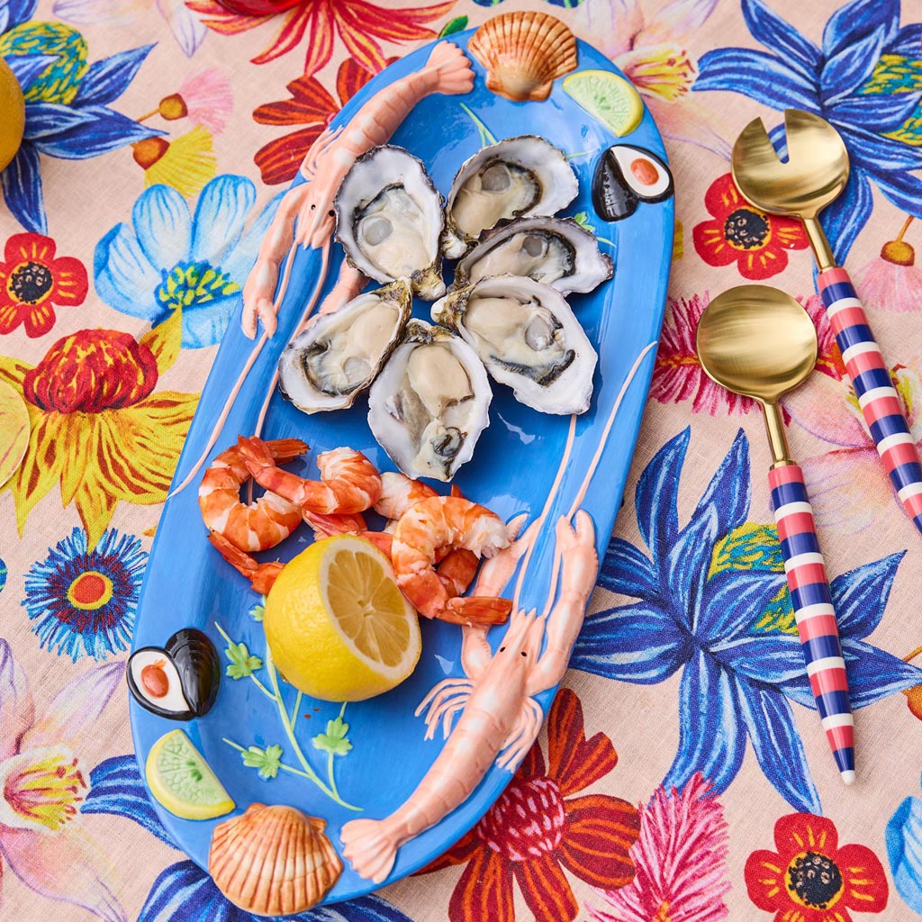 The image is of a large oval platter from Kip & Co, featuring a vibrant and colorful design. The platter is adorned with an artistic depiction of various sea creatures and marine elements, such as fish, shells, and seaweed, all rendered in a playful and whimsical style. The colors are bright and eye-catching, with a mix of blues, greens, pinks, and yellows, creating a lively and cheerful appearance. The platter's design is likely intended to evoke a sense of the ocean and marine life, making it a striking p