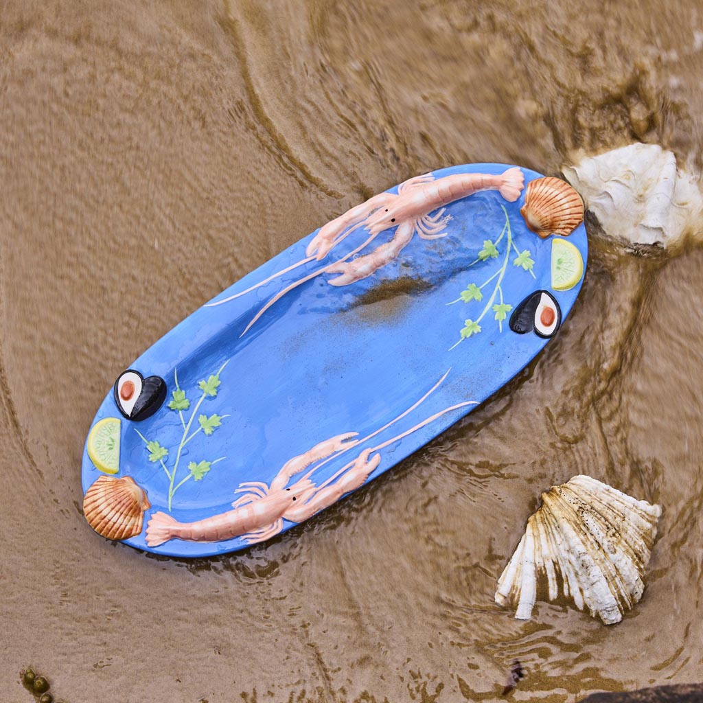 The image is of a large oval platter from Kip & Co, featuring a vibrant and colorful design. The platter is adorned with an artistic depiction of various sea creatures and marine elements, such as fish, shells, and seaweed, all rendered in a playful and whimsical style. The colors are bright and eye-catching, with a mix of blues, greens, pinks, and yellows, creating a lively and cheerful appearance. The platter's design is likely intended to evoke a sense of the ocean and marine life, making it a striking p