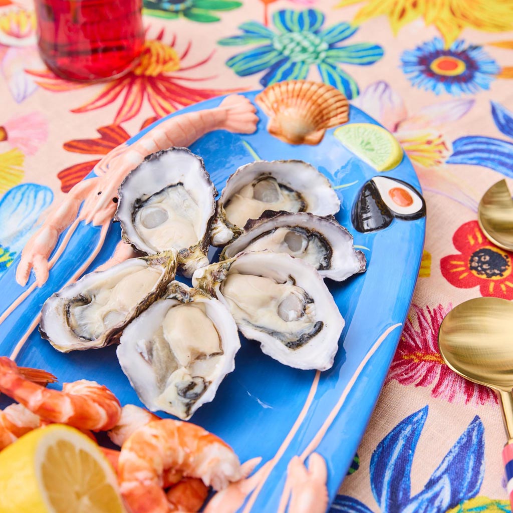 The image is of a small oval platter from Kip & Co, featuring a vibrant and colorful design. The platter is adorned with an artistic depiction of various sea creatures and marine elements, such as fish, shells, and seaweed, all rendered in a playful and whimsical style. The background is a light color, which helps the vivid colors of the marine illustrations stand out. The platter's design is likely intended to evoke a sense of the ocean and coastal life, making it a decorative and functional piece for serv