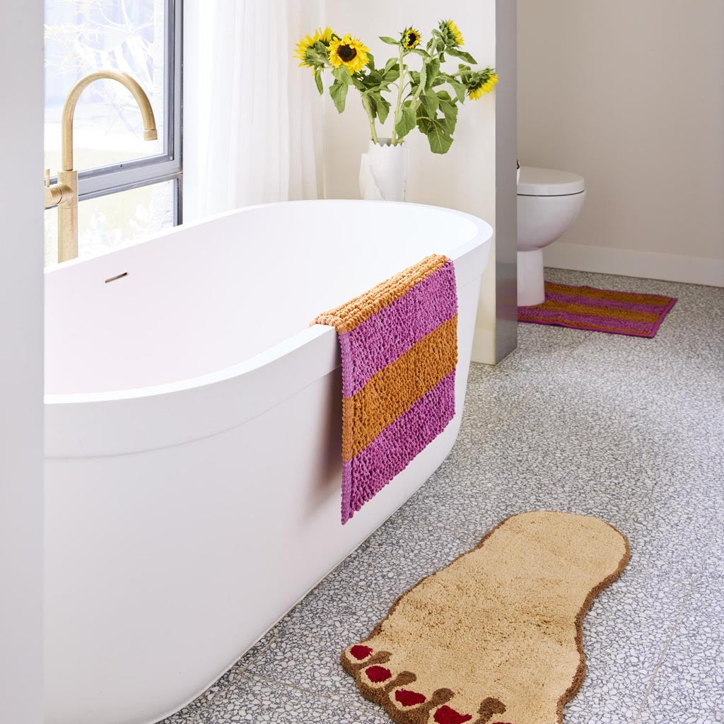 The image is of a colorful bath mat with a striped pattern. The stripes are arranged in a variety of vibrant colors, including shades of pink, orange, yellow, green, blue, and purple. The mat appears to be made of a soft, plush material, likely designed to be both comfortable and absorbent. The overall design is playful and eye-catching, adding a fun and lively touch to a bathroom setting.